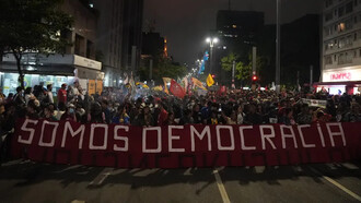 Uma multidão ergue a faixa “somos democracia” durante protesto popular. A disputa entre participação democrática e projetos de poder autoritários está no centro das tensões políticas contemporâneas