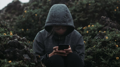 A person sitting on a rock, staring at their phone, surrounded by a sense of grief and the haunting presence of technology