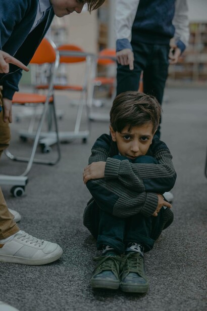 A young boy cowers on the ground in fear as older students surround him, bullying him