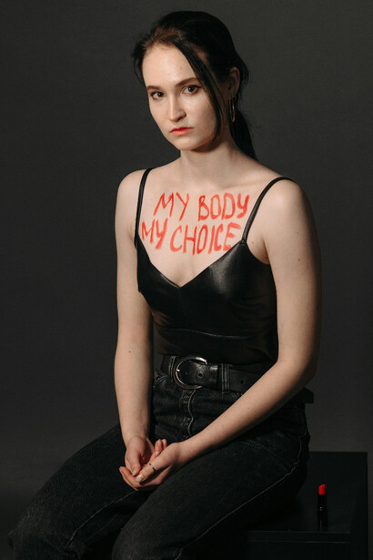 Seated in shadow a figure with “my body my choice” on her chest sits quietly embodying tension and protest