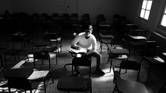 Un estudiante escribe a mano en el interior de un aula prácticamente vacía, rodeado de pupitres desocupados y sin ningún compañero a la vista