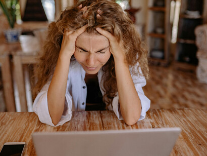 A woman in a white button-up shirt works on her laptop, frustration clearly visible on her face