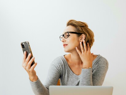 A woman in a gray long-sleeve shirt taking a selfie, frequently glancing at her phone