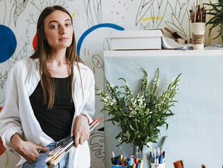 A girl, holding paintbrushes in her hand, gazes dreamily at the camera, surrounded by a large patterned canvas in her home, embodying the artistic workspace and creative process