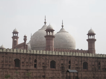 The Badshahi Mosque, glowing in the morning light, stands tall in red and white hues, seen from Minar-e-Pakistan and framed by lush trees