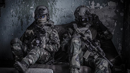 Two officers sit on a the floow, dressed as airsoft players wearing protective glasses, helmets, and holding airsoft guns