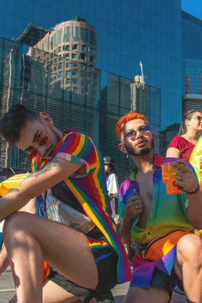 Two individuals pose confidently against the backdrop of a gleaming urban skyline, their vibrant rainbow-themed outfits radiating energy and pride. From bold accessories to striking color coordination, their fashion choices celebrate pride and visibility