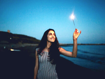 On a dark beach, a woman holds a sparkler, with sparks of light swirling from her fingertips into the night air