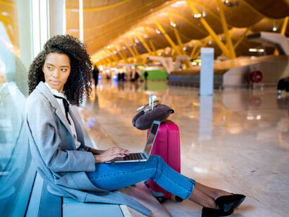 An expat woman works on her laptop while waiting at the airport, seated in front of a large window