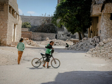 A child stands beside a bicycle amid the ruins of a demolished city in Idlib, Idlib Governorate, Syria