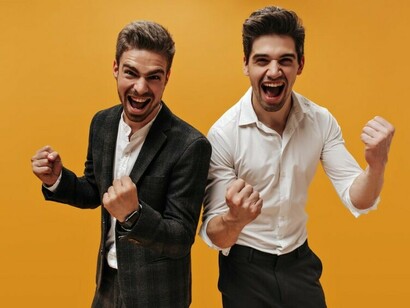 Two stylish young businessmen with dark hair, wearing classic white shirts, share a joyful moment of success against a bright orange backdrop