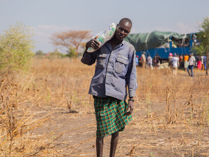 A man transports humanitarian aid in a rural community in South Sudan, with a truck and people visible in the background