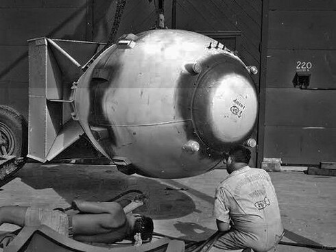Homens trabalham ao lado da Fat Man durante os preparativos para seu emprego, enquanto a estrutura da bomba ocupa o centro da imagem. A cena evidencia como a tecnologia nuclear transformou a capacidade humana de produzir destruição em uma questão de engenharia e logística