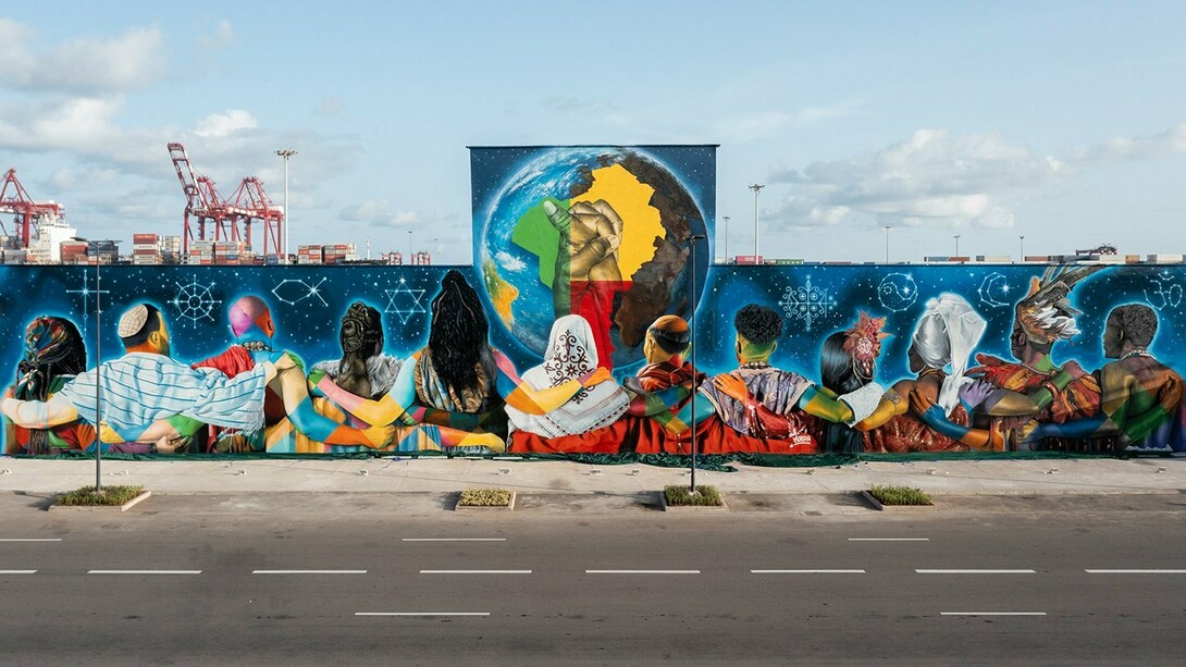 "Coexistence", de Eduardo Kobra, em Cotonou, na República do Benim em 2023. O mural reúne 12 figuras de costas, representando diferentes religiões, unidas diante de um globo que destaca a África e duas mãos entrelaçadas. Realizada na fachada do antigo Hotel do Porto, a obra ocupa cerca de 700 m² e transforma o espaço urbano em uma mensagem visual de tolerância, diálogo inter-religioso e convivência, reafirmando a presença internacional da arte urbana brasileira