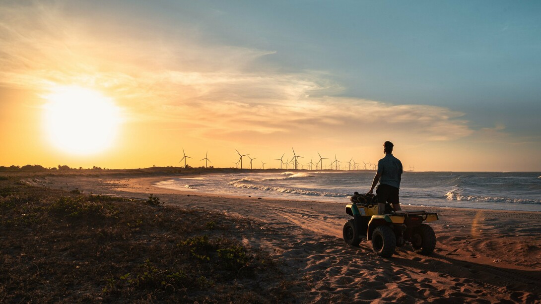 A silhueta de um homem conduzindo um quadriciclo percorre a praia ao entardecer, com aerogeradores ao fundo. A imagem evoca a sensação de liberdade e domínio sobre o ambiente, que o relato contrapõe à necessidade de reconhecer os limites impostos pela natureza