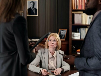 A woman wearing glasses sitting at her desk, listening intently to her subordinates while thinking about her upcoming decisions