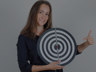 A young woman in a black shirt and denim shorts, pointing hopefully at a dartboard