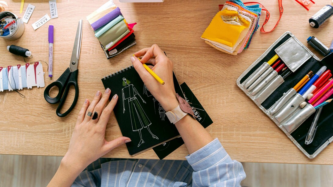 Mãos desenhando roupa em um caderno sobre uma mesa de estudos de moda. Entre traços e ideias, nasce a moda em um processo criativo que une arte, técnica e consciência em cada detalhe. O primeiro esboço de uma peça carrega mais que estética, revela pensamento, cultura e intenção. Antes das passarelas, a moda começa no papel, onde criatividade e propósito se encontram