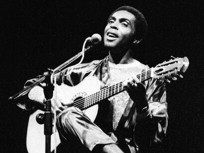 Gilberto Gil no início da carreira, cantando ao microfone e tocando violão em um registro em preto e branco. Muito antes de se tornar presença constante nos palcos que atravessariam a vida do autor, estava começando a construir a trajetória que faria sua música chegar a tantas gerações