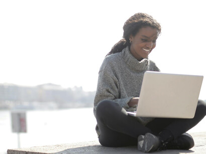 A woman in a gray long-sleeve shirt sits outside in a European city, focused on her laptop as she works remotely