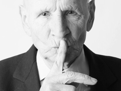An elderly man is firmly pressing his index finger against his lips wishing for silence