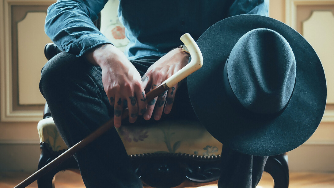 A mafia figure wearing a hat and showing tattoos, holding a cane with a confident stance