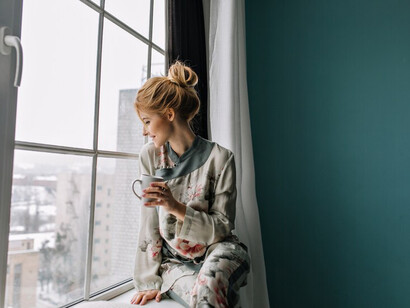 A young blonde woman relaxing with a cup of tea, gazing through a large window, surrounded by the calm ambiance of her home
