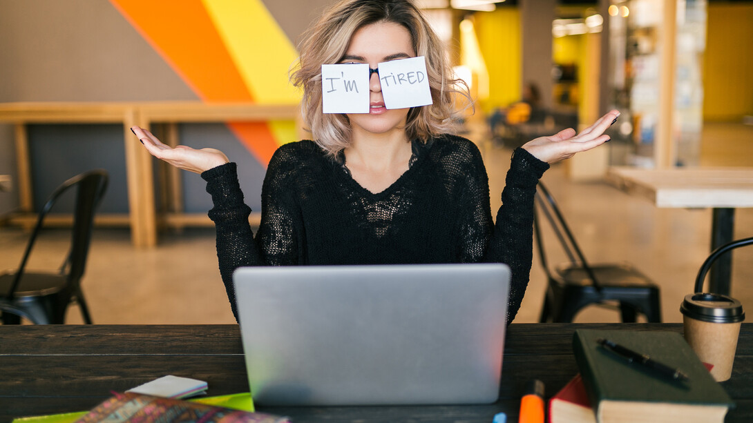 A portrait of a young woman sitting at a table, paper stickers stuck to her glasses, her expression tired and worn