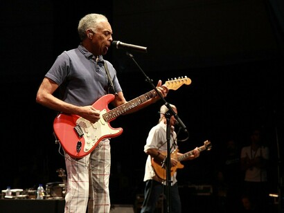 Gilberto Gil canta ao microfone enquanto toca guitarra durante o Festival de Cornouaille, em 2010, em uma apresentação que reúne voz, instrumento e a energia do palco. Para quem acompanhou sua trajetória ao longo de décadas, cada show se transforma também em uma viagem por diferentes épocas da própria vida