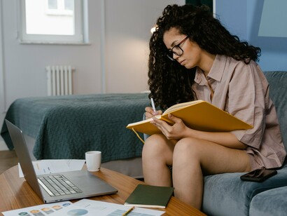 Sentada em um sofá azul, uma mulher escreve em um caderno enquanto mantém o laptop por perto. O papel e a tela aparecem lado a lado, revelando que as práticas tradicionais de leitura e escrita continuam convivendo com os recursos digitais
