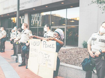 A woman surrounded by police officers is holding a cardboard sign which reads "world peace no more violence. It starts with you" 