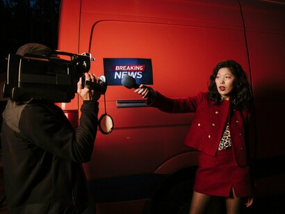 A journalist reporting live as a photographer frames the shot beside a news van, reflecting the pursuit of compelling visuals and headlines