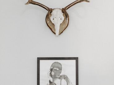 Parede branca com um quadro de moldura preta se destaca. Acima do quadro, os chifres de um animal exibido como se fosse arte