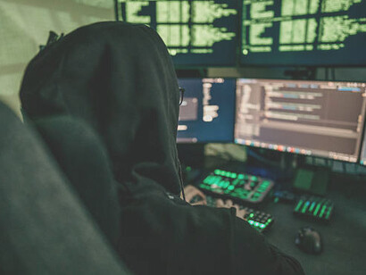 A hacker coding on multiple computer screens in a dark room, representing cyber warfare and emerging cyber threats
