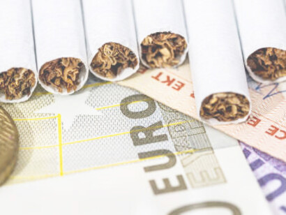 Cigarettes alongside the word 'TAX' on a dark background, with Euro banknotes in the backdrop
