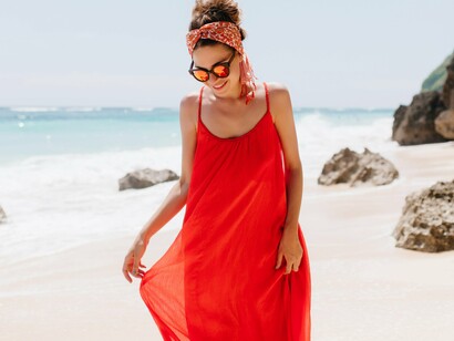 A person dressed in a long, flowing red dress stands on a sandy beach, possibly enjoying a warm, coastal setting, suggesting a preference for practical yet stylish clothing suitable for a relaxed environment