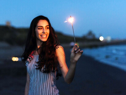 A woman on a moonlit beach, holding a sparkler as streams of glowing energy radiate from her fingertips