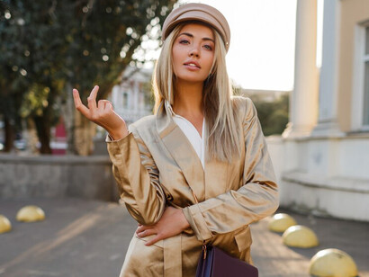 A fashionable blonde woman in a beige cap and jacket walks confidently through the autumn streets, illuminated by the soft glow of sunset, showcasing timeless style