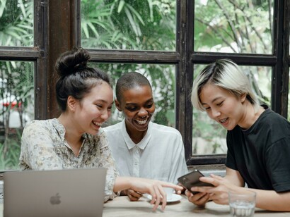 Smiling multiethnic women enjoying conversation and browsing on their devices in a café, illustrating the social nature of apps and digital platforms