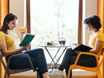 Due giovani donne che leggono al tavolino di un locale pubblico: "... il linguaggio non è mai solo uno scambio di dati: è relazione, contesto, emozione, trasformazione reciproca"