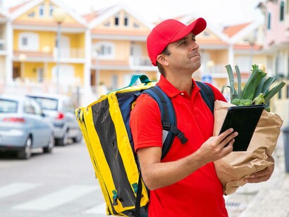 Fattorino con zaino termico e cappellino rosso consulta un tablet tenendo una busta della spesa contenente verdura fresca, in una via cittadina