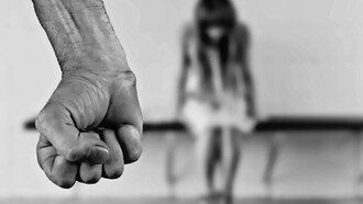 A man stands with his fists clenched in anger, while a woman sits on the ground in front of him, fearful and covering her face