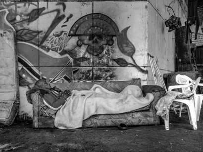 A homeless man lies on a sofa on the street, with vibrant graffiti decorating the wall behind him