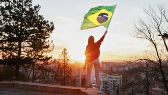 Uma mulher de calça branca e camisa vermelha segura a bandeira brasileira diante de uma cidade iluminada pelo sol. Mais do que um símbolo nacional, a cena sugere a presença feminina como parte ativa das escolhas que definem os caminhos do Brasil