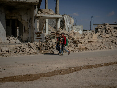 Two students walk along a dusty, unpaved road in Idlib, Syria, passing through the shattered remains of a city devastated by conflict