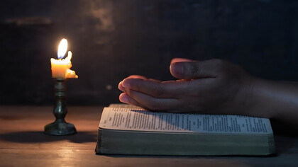 The woman's hand praying on a table upon a Bible with a candle next to it