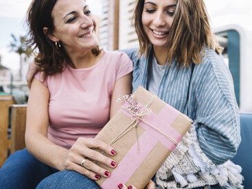 A woman surprises her mother with a gift and a warm hug on the sofa for Mother’s Day