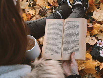 A person reading a book and holding a cup of coffee, surrounded by autumn leaves
