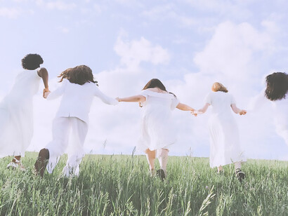 Five women running hand in hand in white dresses on the grass in nature