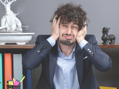 A burned-out and agitated businessman is depicted sitting at his desk, visibly holding his head, showcasing the effects of stress and burnout in the workplace